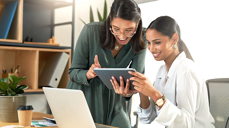 Two women in an office smiling while looking at a tablet