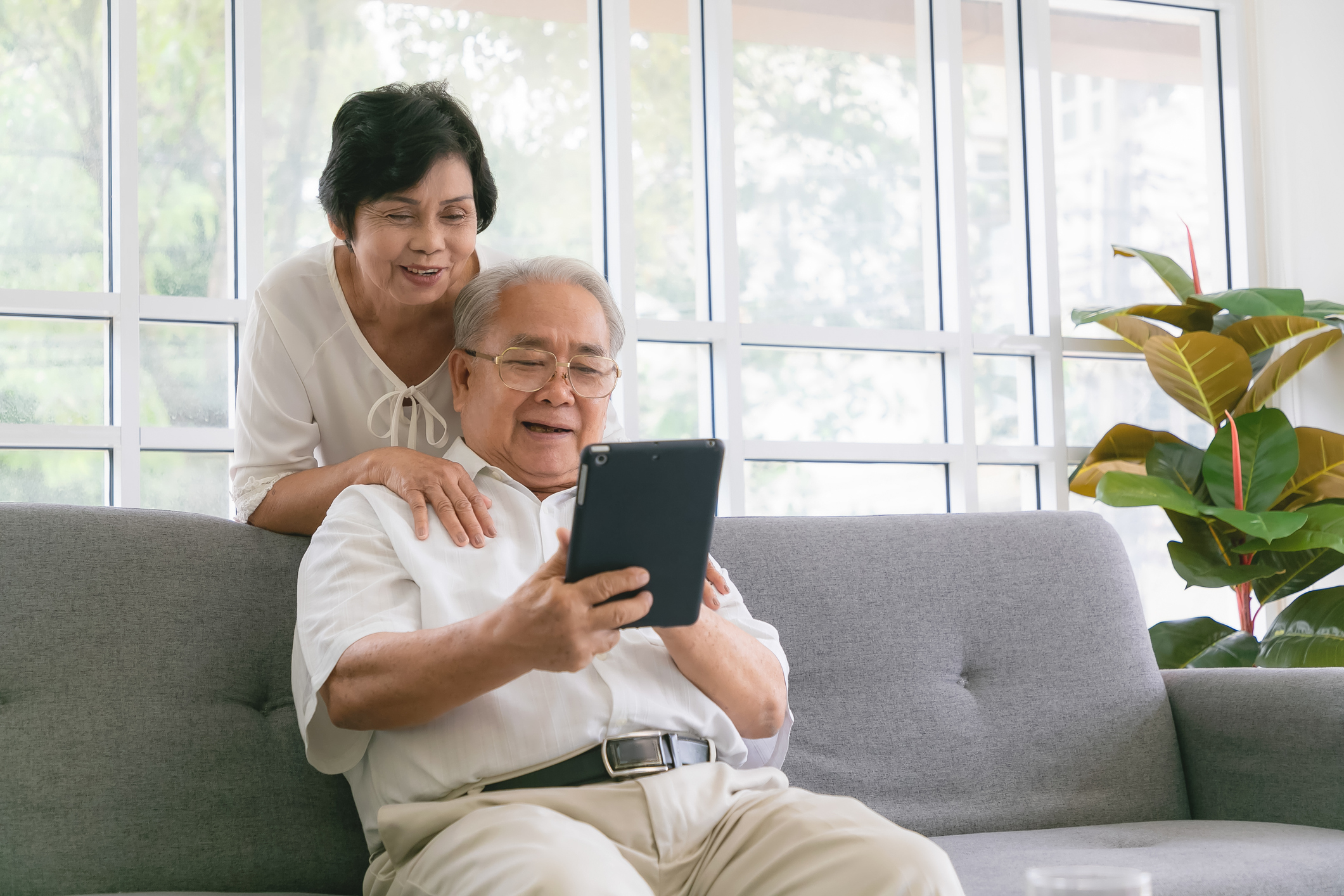 Couple of seniors smiling and looking at the same tablet hugged on the sofa.